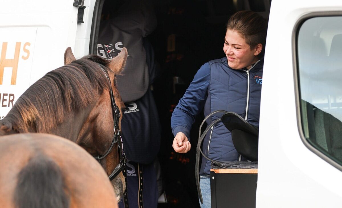 Zadelpasser in de bus met een zadel op de werkbank, terwijl een paard naast de bus staat en contact maakt.